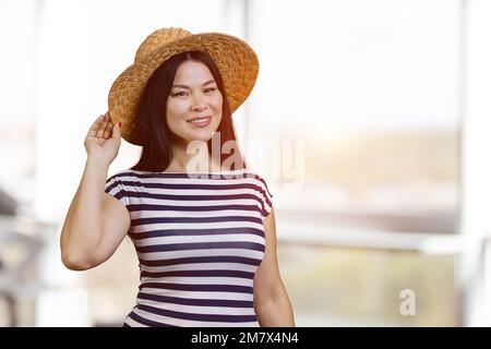 Bonne jeune femme asiatique portant un chapeau de paille à l'intérieur. Arrière-plan de fenêtres flou. Banque D'Images