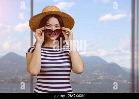 Portrait d'une jeune femme asiatique en chapeau de paille et chemise rayée met sur des lunettes de soleil. Paysage de montagne de l'intérieur. Banque D'Images