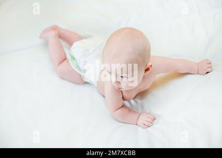 Un petit enfant se lève la tête sur du sang blanc. Un bébé heureux apprend à ramper et à grimper sur les poignées. Prendre soin d'un enfant dans une couche. Banque D'Images