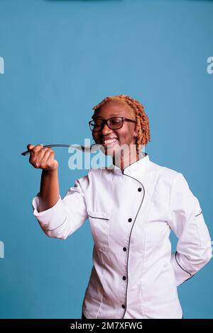 Un professionnel souriant portant un blouson de chef teste une nouvelle recette avec une cuillère en studio. Chef afro-américain optimiste en uniforme, enthousiaste à l'idée de préparer un nouveau plat sur fond bleu. Banque D'Images