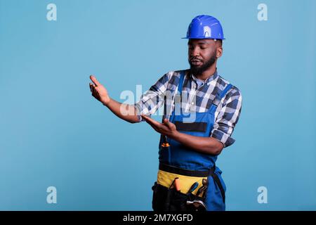 Travailleur de la construction portant un casque de sécurité prétendant tenir la bannière publicitaire dans le studio tourné sur fond bleu. Constructeur avec les bras levés comme si porter quelque chose et regarder attentivement. Banque D'Images