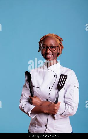 Chef de cuisine afro-américaine du restaurant tenant des ustensiles de cuisine dans une prise de vue en studio. Femme membre du personnel de cuisine avec des cheveux bouclés et une veste uniforme souriant sur fond bleu. Banque D'Images