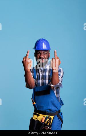 Un constructeur américain africain agacé envoyant tout à l'enfer fatigué du travail excessif. Faire des panneaux de désapprobation avec les deux mains regardant dans le dégoût de la caméra sur un fond bleu. Banque D'Images