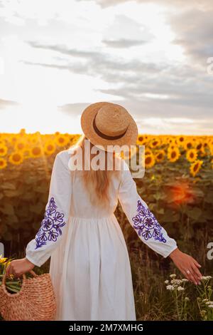 Bonne femme marchant dans le champ de tournesol en fleur au coucher du soleil tenant sac avec des fleurs. Paysage d'été Banque D'Images