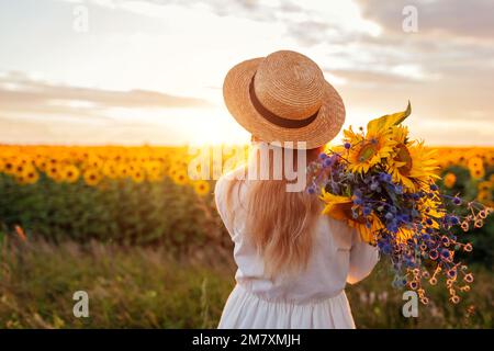 Vue arrière de la femme appréciant la vue dans le champ de tournesol en fleur au coucher du soleil avec bouquet de fleurs. Paix et liberté Banque D'Images