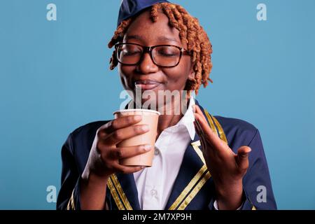 Une femme professionnelle se réjouit de l'odeur du café fraîchement préparé pendant la pause au travail. Hôtesse de l'air vêtue d'un uniforme de travail sur fond bleu. Banque D'Images