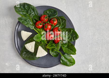 Tomates cerises rouges sur une branche, feuilles d'épinards verts et fromage mozzarella sur une plaque sombre sur une table en béton clair Banque D'Images