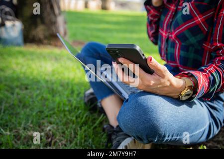 Gros plan sur les mains d'une fille en utilisant son smartphone et son ordinateur portable assis dans un parc public Banque D'Images