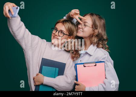 Portrait de deux jeunes filles heureuses tenant des copybooks et prenant un selfie isolé sur fond vert. Prise de selfie, préparation aux examens. Banque D'Images