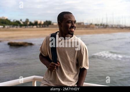 Jeune Afro-américain en vêtements décontractés avec sac à dos debout sur un remblai près de la mer et en regardant la plage Banque D'Images