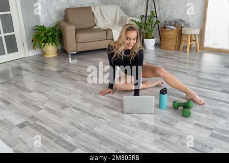 Jeune entraîneur blond yoga instructeur en utilisant la tablette enseigne le marathon de forme physique à distance, tutoriel en ligne. Femme athlétique s'étirant Banque D'Images