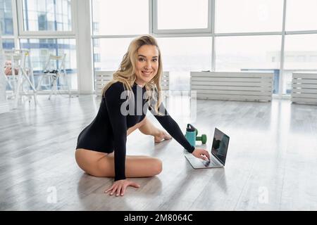 Femme sportive blonde assise sur le sol à la maison, enseigne le marathon de fitness à distance. Jeune formateur, instructeur, formation à l'aide d'un ordinateur portable. Sportif Banque D'Images