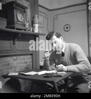 1950s, historique, un homme portant une chemise et une cravate et un chandail assis à une petite table près d'une cheminée dans la chambre à l'avant à la maison, fumant son pipe et écrivant une lettre, Angleterre, Royaume-Uni. Banque D'Images