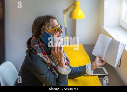 une femme avec des lunettes jaunes parle au téléphone dans son bureau tout en tenant un ordre du jour dans sa main. elle sourit. télétravail et concept indépendant Banque D'Images