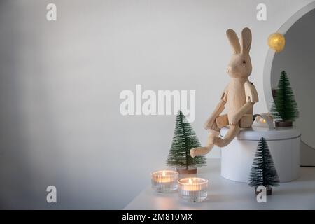 Un lièvre en bois articulé, un petit sapin de Noël vert artificiel et des bougies allumées en chandeliers sur une commode blanche. Espace pour le texte. Christm Banque D'Images