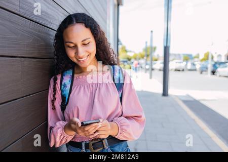 Femme étudiante d'université noire souriante, debout à côté d'un mur en bois et tenant un mobile. Banque D'Images