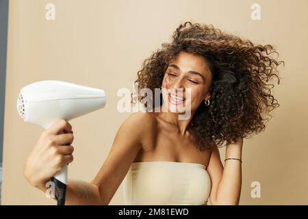 Femme sèche les cheveux freux avec sèche-cheveux, maison beauté produits coiffants cheveux, sourire sur fond beige Banque D'Images