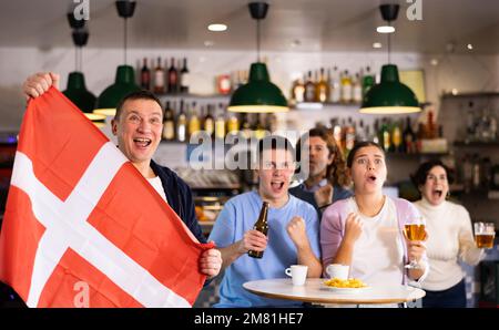 Compagnie de jeunes adultes fans de sport soutenant l'équipe danoise avec drapeau d'état tout en buvant de la bière au bar Banque D'Images