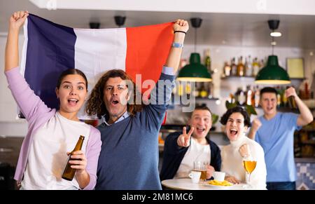 Compagnie de jeunes fans sportifs enthousiastes pour adultes qui agite le drapeau de la France et soutient l'équipe nationale avec de la bière et des en-cas au pub Banque D'Images