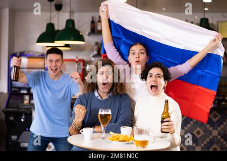 Groupe de jeunes amis adultes enthousiastes qui agite le drapeau de la Russie et qui soutiennent l'équipe nationale avec de la bière au bar Banque D'Images