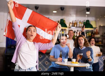 Compagnie de jeunes adultes fans de sport soutenant l'équipe danoise avec drapeau d'état tout en buvant de la bière au bar Banque D'Images