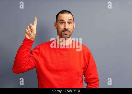 Portrait d'un homme hispanique barbu vêtu d'un pull-over rouge heureux excité pointant le doigt vers l'espace publicitaire isolé sur fond gris. Banque D'Images