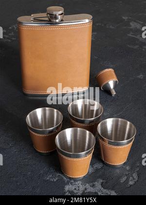 Flasque de hanche en acier inoxydable et cuir brun avec tasses sur fond sombre, foyer sélectif. Concept de boissons alcoolisées, boissons alcoolisées Banque D'Images