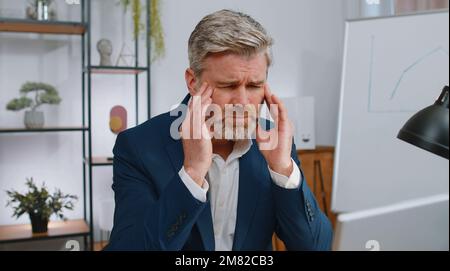 Homme d'affaires fatigué et mûr souffrant de maux de tête problèmes de tension et de migraine, stress à la maison bureau lieu de travail. Un travailleur indépendant d'âge moyen travaille sur un ordinateur portable. Travail excessif Banque D'Images