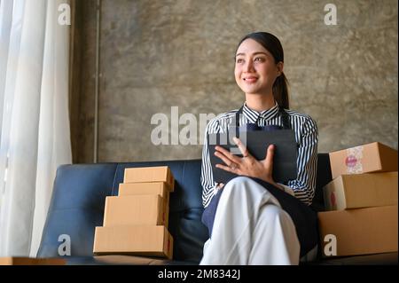 Attrayante et charmante jeune femme asiatique vendeur en ligne ou entrepreneur en commerce électronique utilisant une tablette numérique sur un canapé dans son salon. Banque D'Images