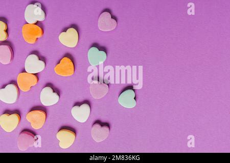 Bonbons à base de sucre en forme de cœur coloré éparpillés sur fond violet Banque D'Images