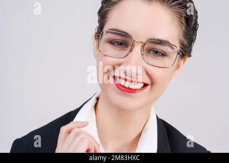 Gros plan portrait jeune femme d'affaires qui semble heureuse et confiante. Souriez sur son visage, dans un costume noir et une chemise blanche avec une courte coupe de cheveux avec rouge à lèvres sur un fond gris. Banque D'Images