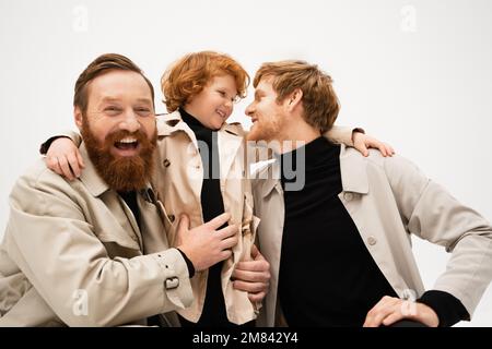 joyeux barbu homme en train de rire à la caméra près de redhead fils et petit-fils dans des manteaux de tranchée isolés sur gris, image de stock Banque D'Images