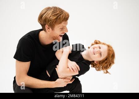 homme excité en t-shirt noir jouant avec heureux fils à tête rouge croisant les bras isolés sur gris, image de stock Banque D'Images