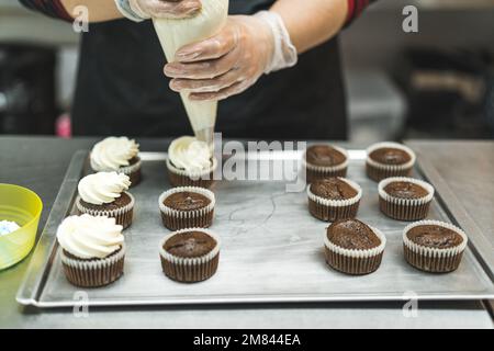 gros plan des mains de femmes avec des gants de crème sur des cupcakes au chocolat, boulangerie. Photo de haute qualité Banque D'Images