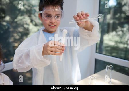 Détails tube à essai avec sortie de solution dans les mains d'un garçon d'école en blouse de laboratoire et lunettes de protection, conduite d'expérience chimique Banque D'Images