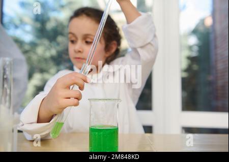 Détails: Pipette graduée et tube à essai avec liquide chimique dans les mains d'une écolière primaire floue. Classe de chimie Banque D'Images