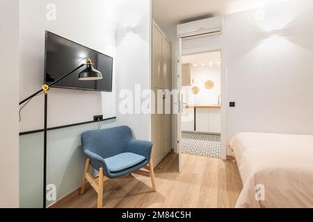 Petite chambre confortable avec lit recouvert d'une couverture beige. Confortable fauteuil bleu dans lequel vous pouvez lire par la lumière de la veilleuse. Grand téléviseur sur le mur opposé Banque D'Images