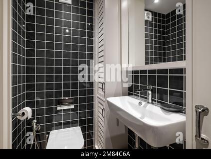 Belle salle de bains avec carreaux carrés noirs brillants, lavabo en céramique blanche avec robinet et casiers en bois pour les serviettes. Le rétroviseur reflète la partie supérieure de Banque D'Images