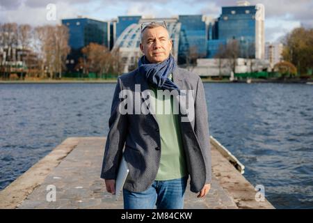 Homme d'affaires heureux de 50 ans dans des vêtements élégants près de l'étang d'eau avec ordinateur portable. Homme marche dehors dans le parc de la ville d'automne. Banque D'Images