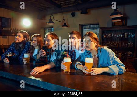 Groupe de jeunes, hommes et femmes, fans de sport regardant le match au pub. Regarder attentivement, encourager émotionnellement l'équipe favorite. Banque D'Images