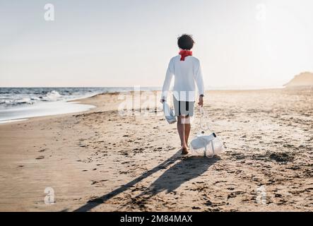 vue arrière d'un garçon dans la distance collectant des conteneurs en plastique sur la plage. il porte un sac plein de conteneurs. concept d'écologie et de soin Banque D'Images