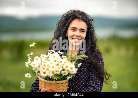 Une femme se tient sur un champ vert et tient un panier avec un grand bouquet de pâquerettes entre ses mains. En arrière-plan, il y a des montagnes et un lac. Banque D'Images