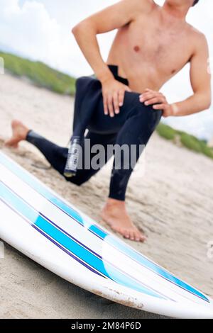 Préparation pour frapper les vagues. Un jeune surfeur s'étire sur la plage à côté de son conseil. Banque D'Images