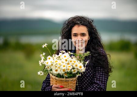 Une femme se tient sur un champ vert et tient un panier avec un grand bouquet de pâquerettes entre ses mains. En arrière-plan, il y a des montagnes et un lac. Banque D'Images