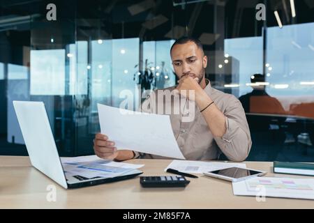 Un homme d'affaires sérieux et péneux lisant un rapport financier, un homme d'affaires hispanique tenant un document entre les mains et regardant déçu, travaillant dans un bureau moderne avec un ordinateur portable derrière un travail papier. Banque D'Images