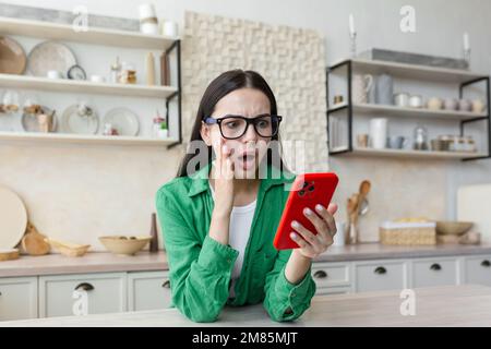 Une jeune femme inquiète est debout à la maison dans la cuisine, tenant le téléphone dans ses mains. Choqué elle lit les nouvelles, a reçu un message, vérifie son compte. Banque D'Images