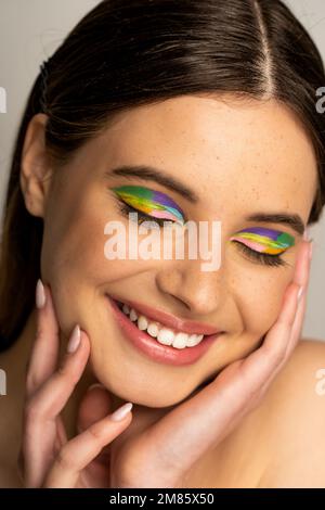 Heureuse fille de l'adolescence avec maquillage coloré touchant le visage isolé sur gris Banque D'Images