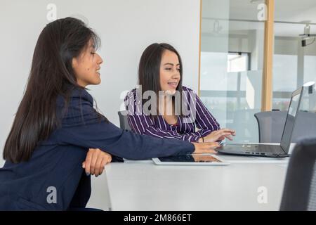 Deux collègues de travail discutent tout en examinant un projet sur leur ordinateur portable. Femmes latines travaillant au bureau. Banque D'Images