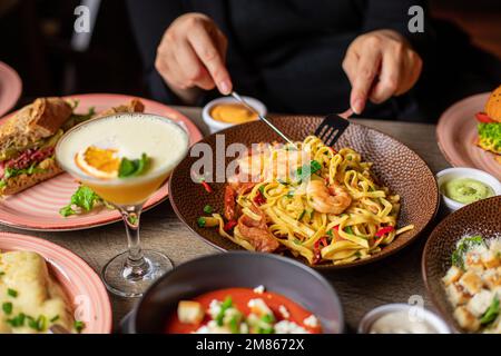 L'homme à la fourchette et au couteau mange des pâtes avec des crevettes de l'assiette sur la table pleine de nourriture délicieuse. Plats chauds et boissons alcoolisées pour le dîner de fête Banque D'Images