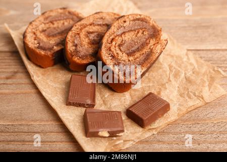 Papier à pâtisserie avec morceaux de délicieux rouleaux de gâteau éponge et chocolat sur table en bois Banque D'Images